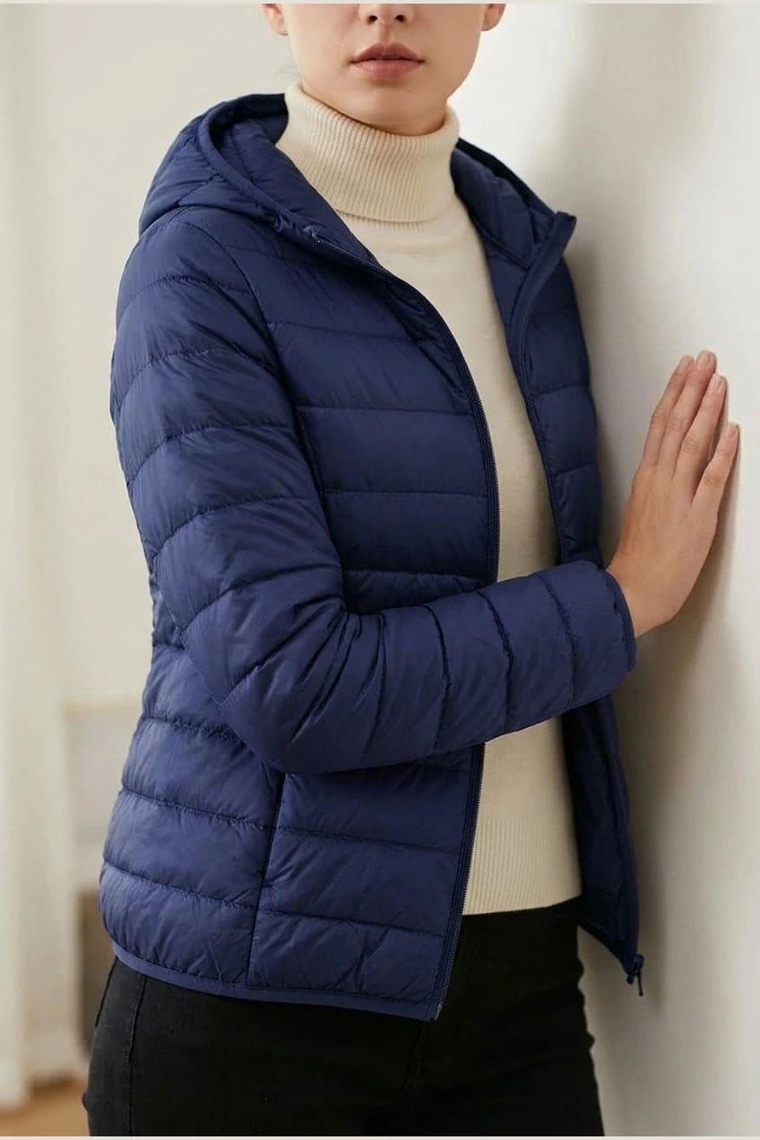 Kvinde i en let quiltet hættejakke med lynlås, båret åben over en rullekravesweater og mørke bukser._1
