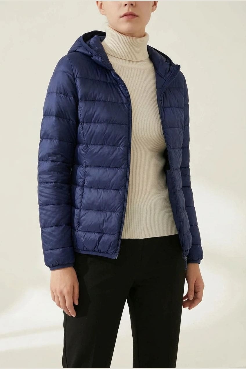 Kvinde i en let quiltet hættejakke med lynlås, båret åben over en rullekravesweater og mørke bukser._3