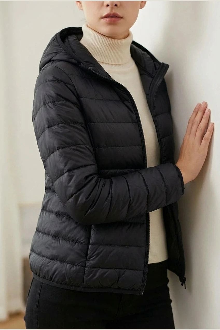 Kvinde i en let quiltet hættejakke med lynlås, båret åben over en rullekravesweater og mørke bukser._6