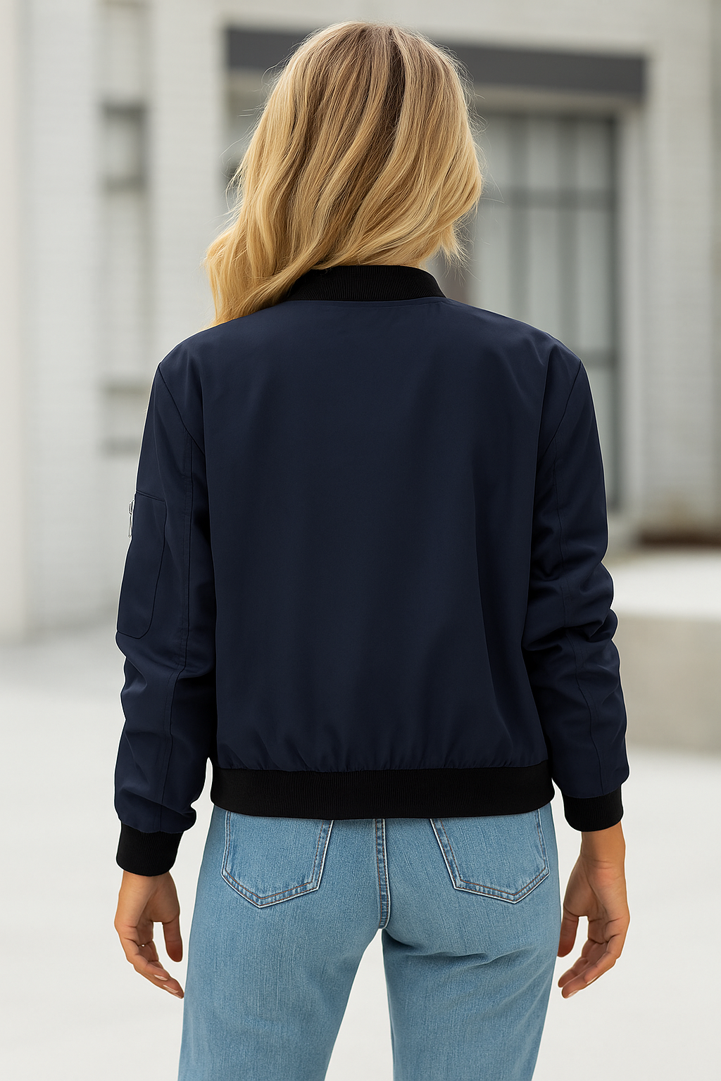 Kvinde iført en mørk bomber jakke med lynlås, hvid top og jeans, holder en kaffekop udendørs._1