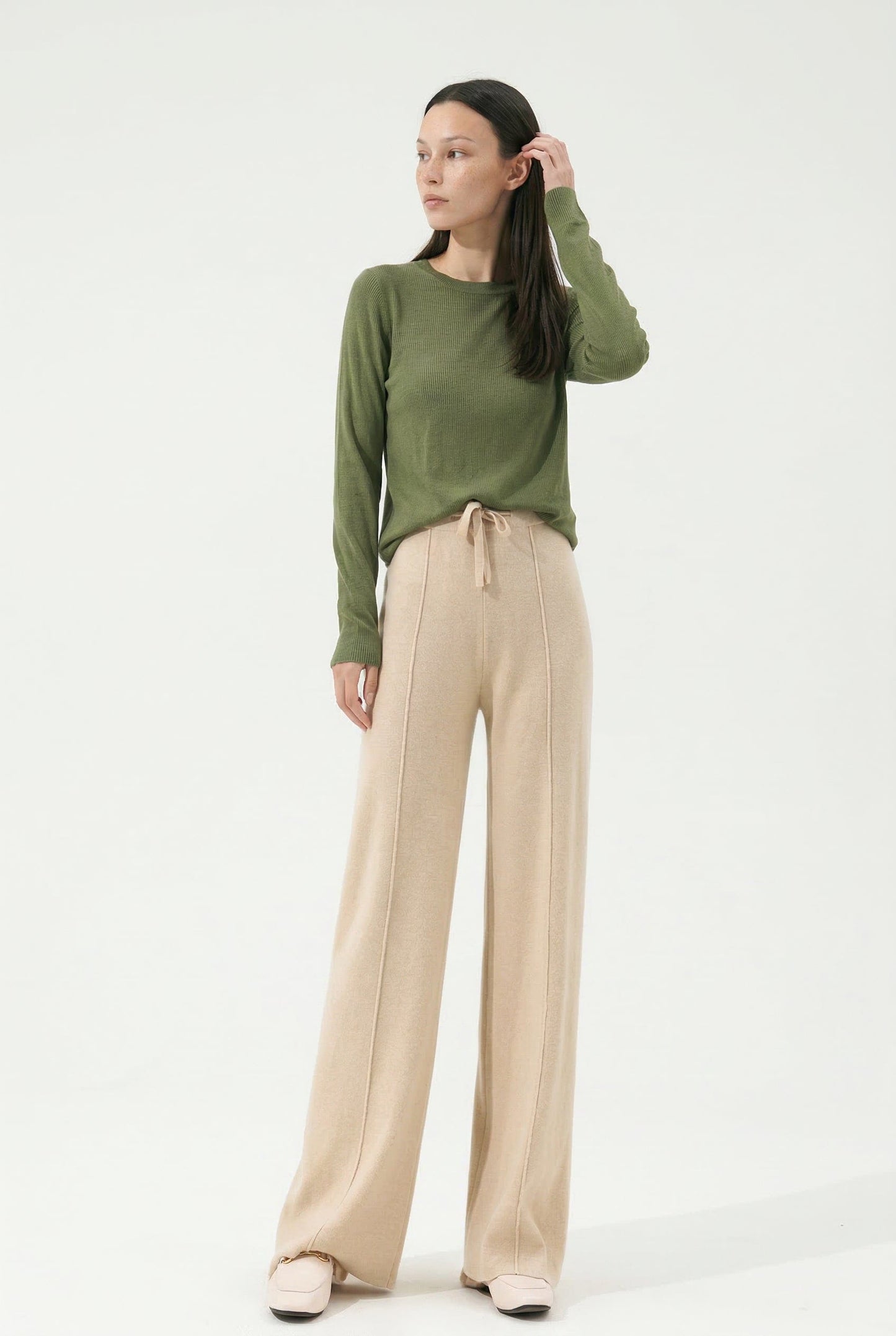 Kvinde iført olivengrøn sweater og beige vide bukser med snørelukning i taljen, stående i naturligt lys._7