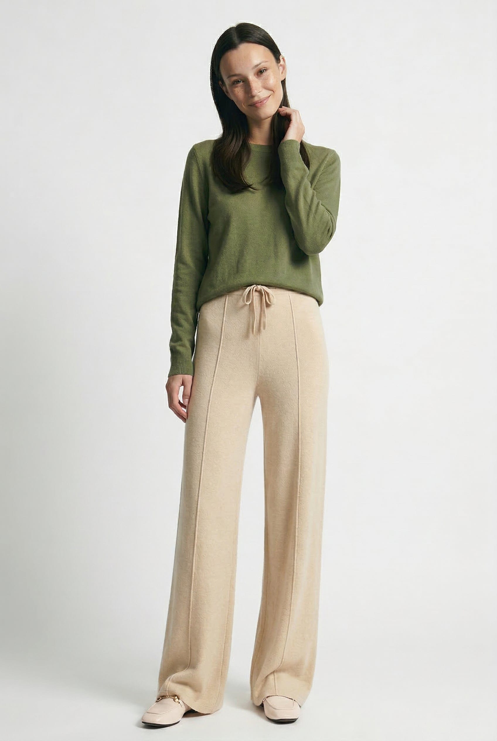 Kvinde iført olivengrøn sweater og beige vide bukser med snørelukning i taljen, stående i naturligt lys._8