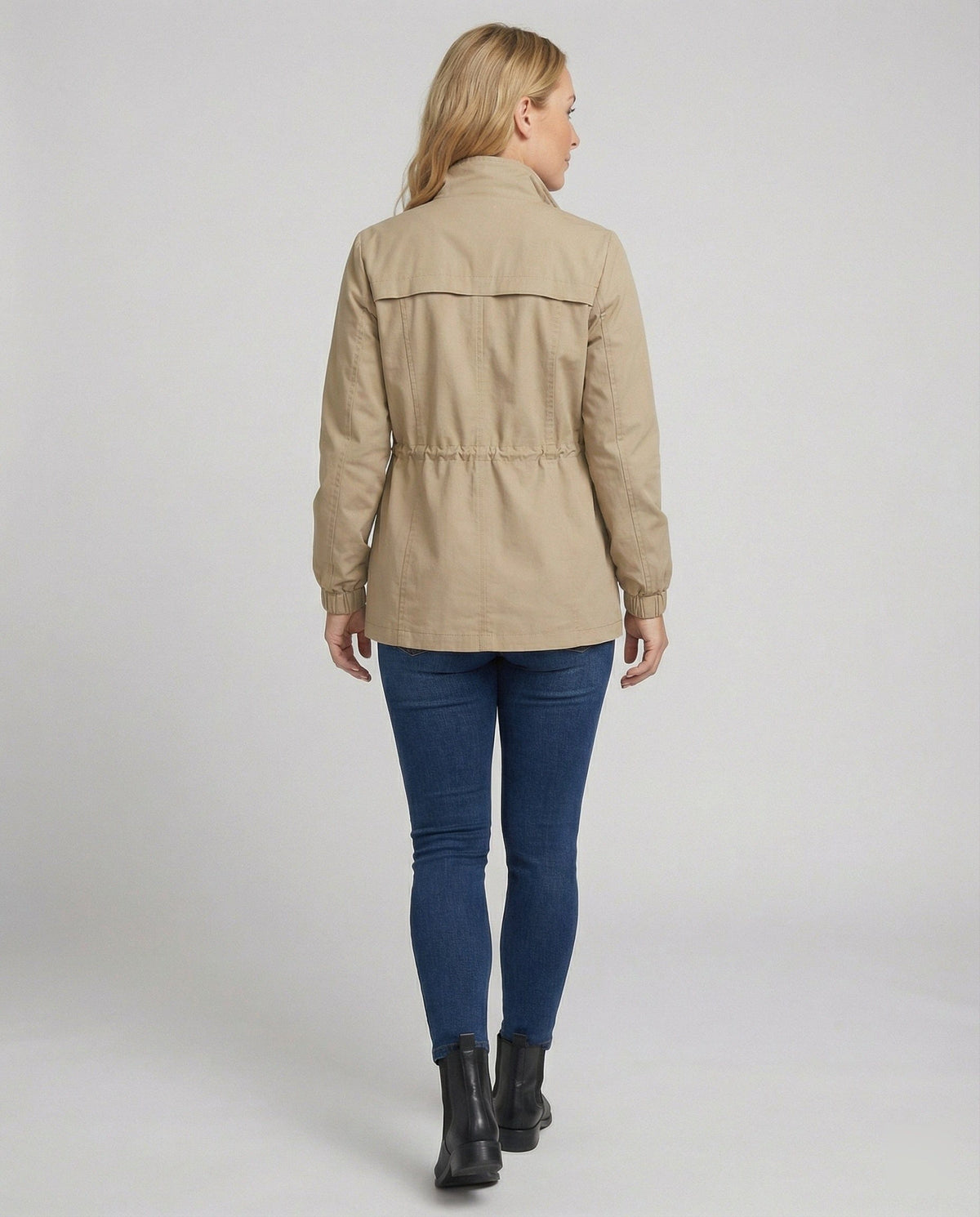 Kvinde i lang utility-jakke med høj krave, lynlås, snøre i taljen og store lommer, styret med jeans og hvid T-shirt._1