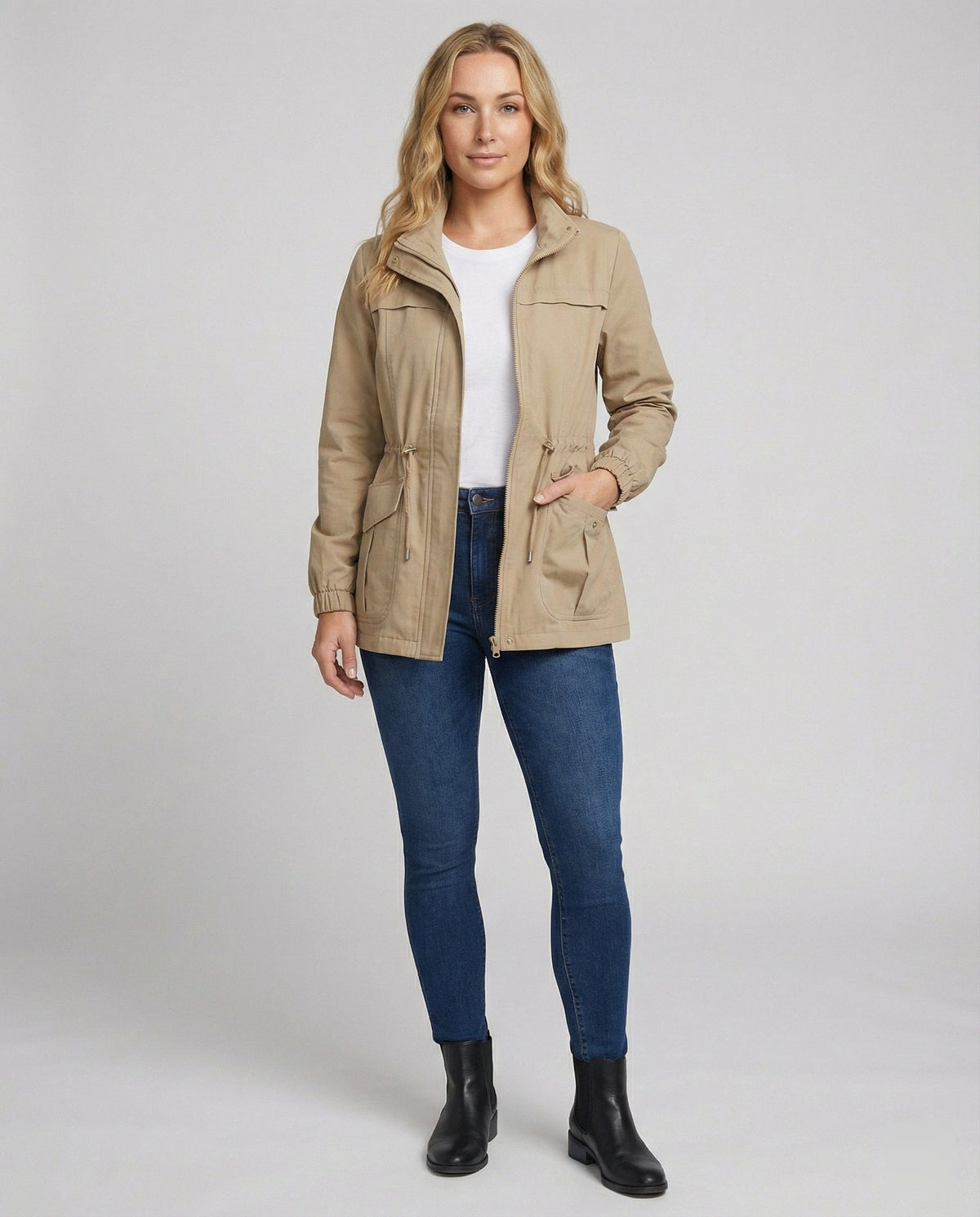 Kvinde i lang utility-jakke med høj krave, lynlås, snøre i taljen og store lommer, styret med jeans og hvid T-shirt._2