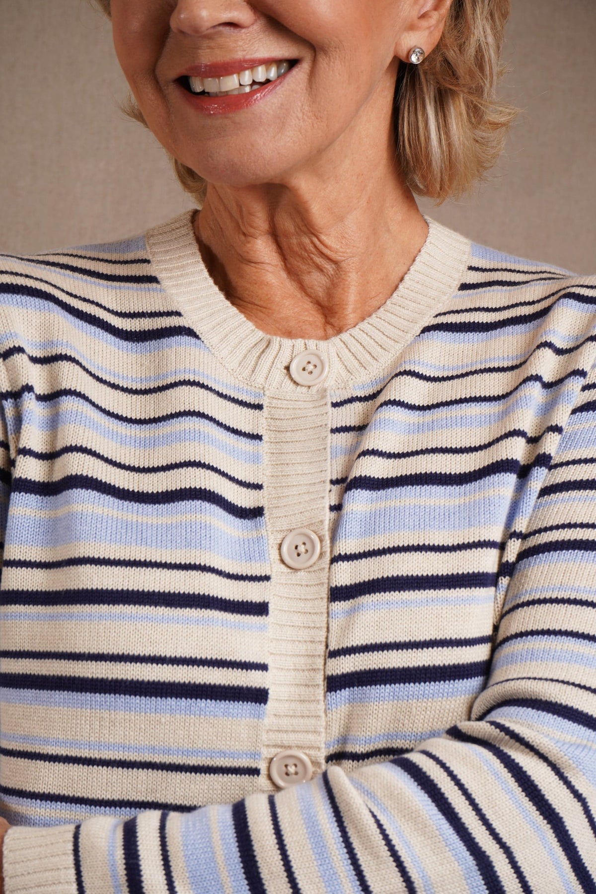 Kvinde iført åben stribet strikcardigan med knapper og rund hals over en hvid T-shirt og lange bukser, stående ved en træportlåge._2