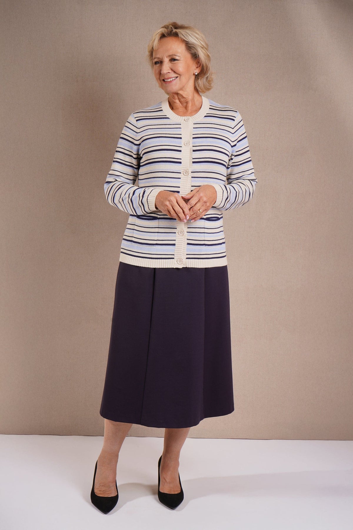 Kvinde iført åben stribet strikcardigan med knapper og rund hals over en hvid T-shirt og lange bukser, stående ved en træportlåge._4