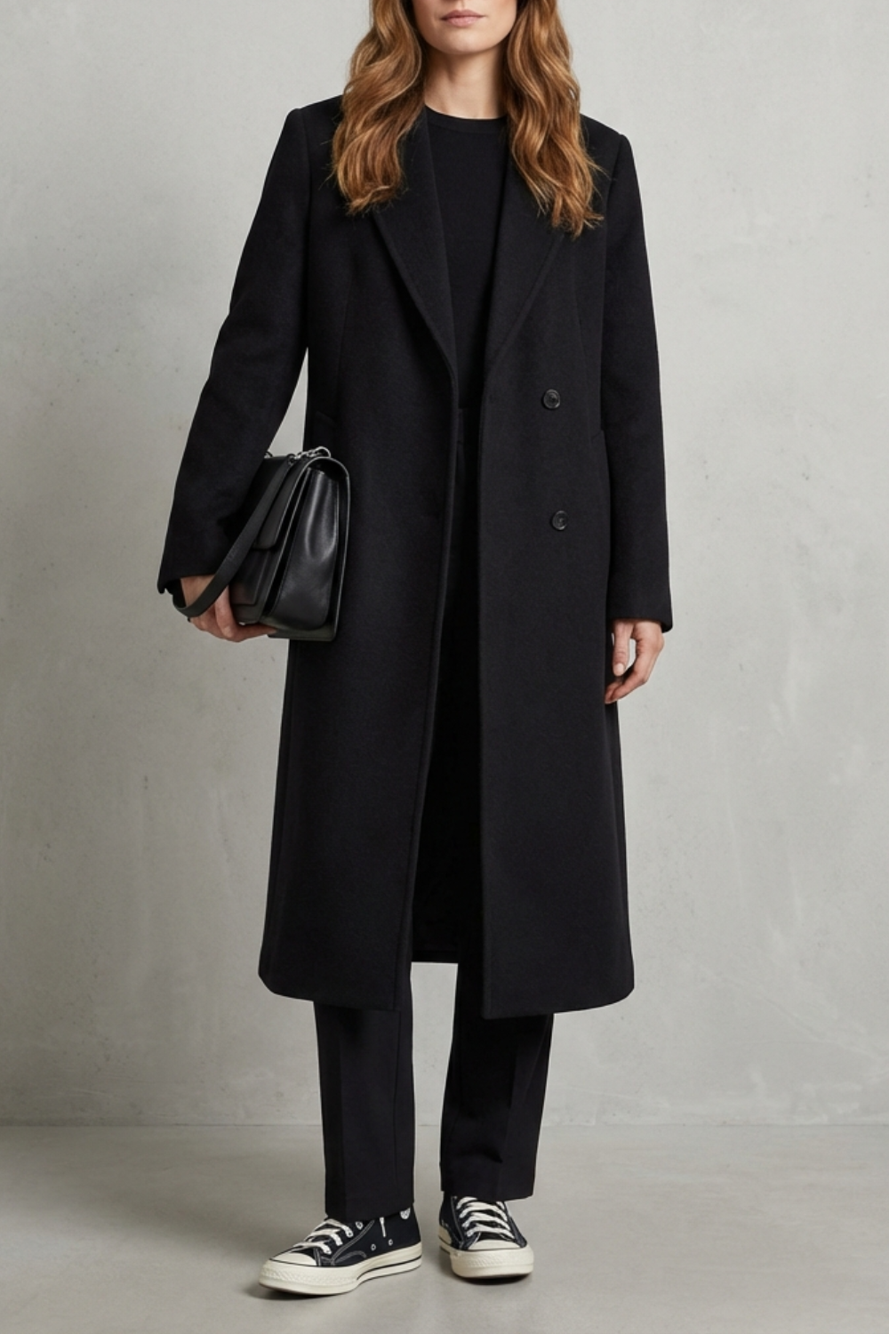 Kvinde iført en lang sort dobbeltradet overcoat med brede revers, sort taske og sneakers, fotograferet mod en neutral baggrund._0