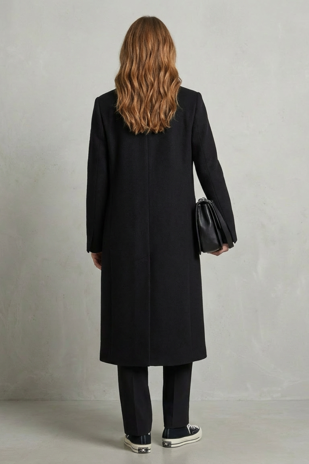 Kvinde iført en lang sort dobbeltradet overcoat med brede revers, sort taske og sneakers, fotograferet mod en neutral baggrund._3