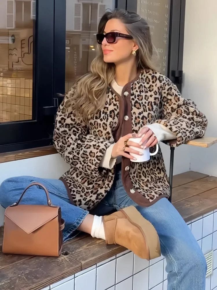 Kvinde iført leopardmønstret cardigan med brunt kontrastpanel og knaplukning over jeans og støvler, siddende på en bænk udenfor en café_2