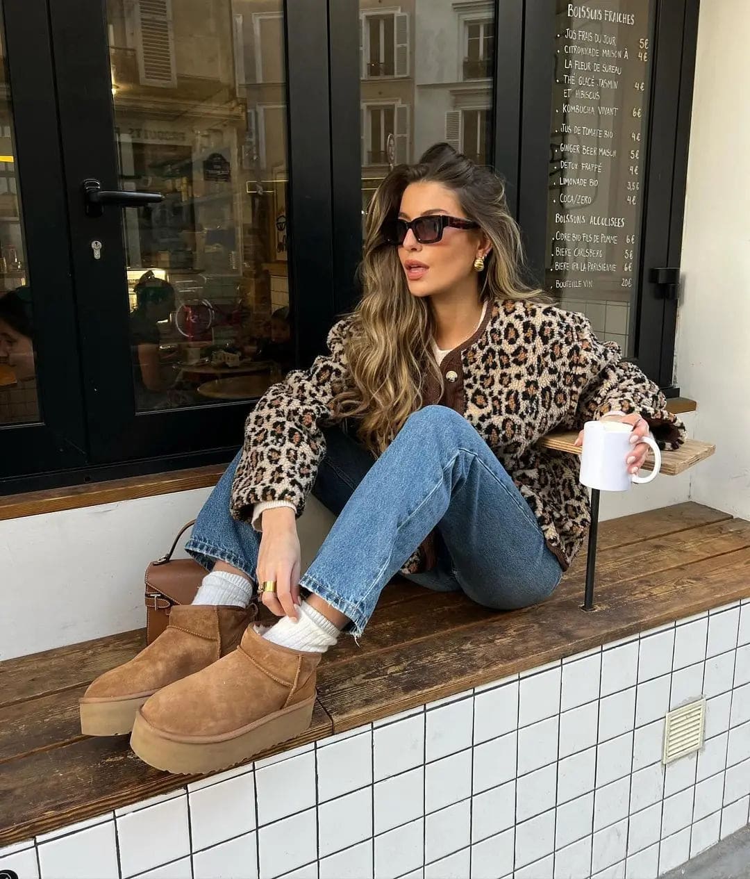 Kvinde iført leopardmønstret cardigan med brunt kontrastpanel og knaplukning over jeans og støvler, siddende på en bænk udenfor en café_3
