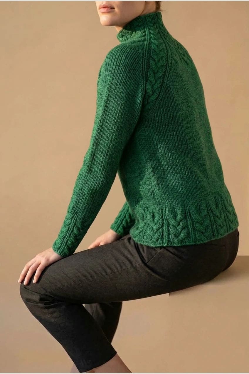 Kvinde i strikket sweater med høj hals, raglanærmer og snoningsmønster ved skuldre og nederste kant._11