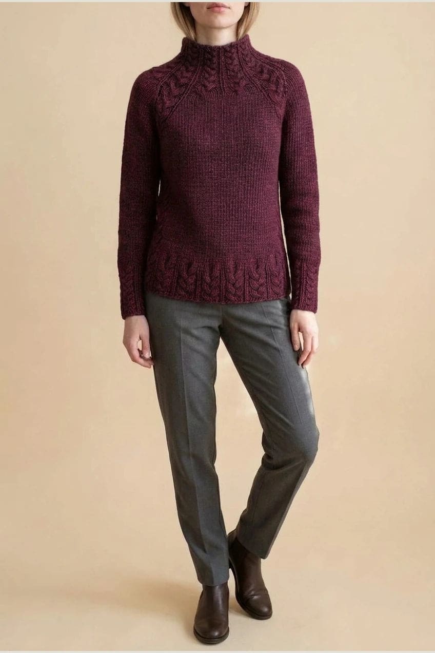 Kvinde i strikket sweater med høj hals, raglanærmer og snoningsmønster ved skuldre og nederste kant._19