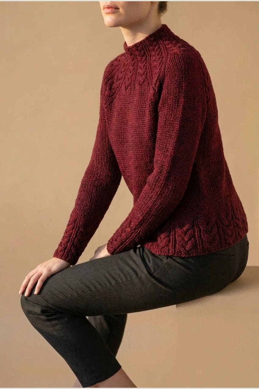 Kvinde i strikket sweater med høj hals, raglanærmer og snoningsmønster ved skuldre og nederste kant._23