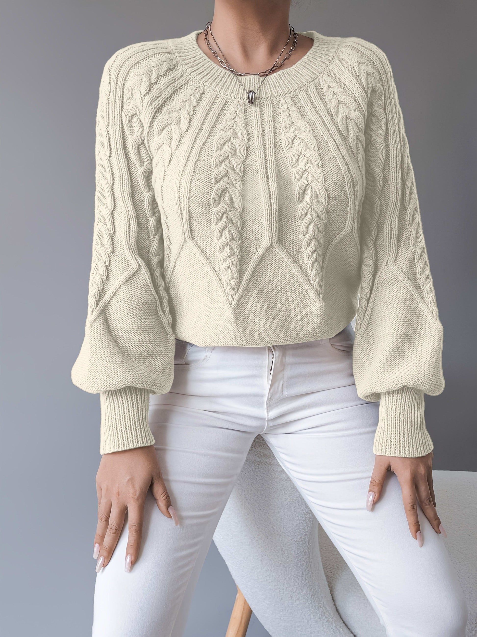 Kvinde iført sort strikket sweater med kabelstrik og ballonærmer, stylet med hvide bukser og halskæde, fotograferet forfra_2
