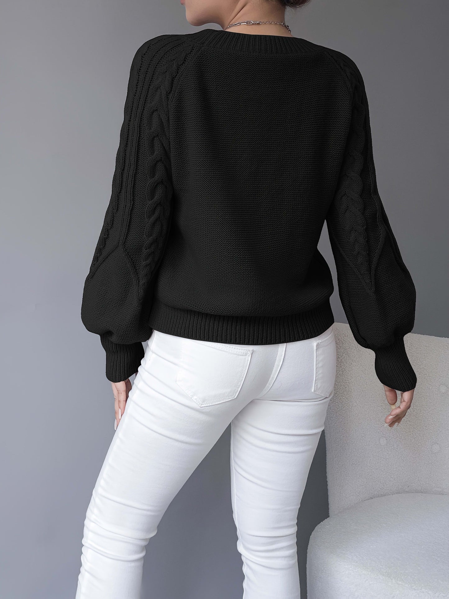 Kvinde iført sort strikket sweater med kabelstrik og ballonærmer, stylet med hvide bukser og halskæde, fotograferet forfra_5