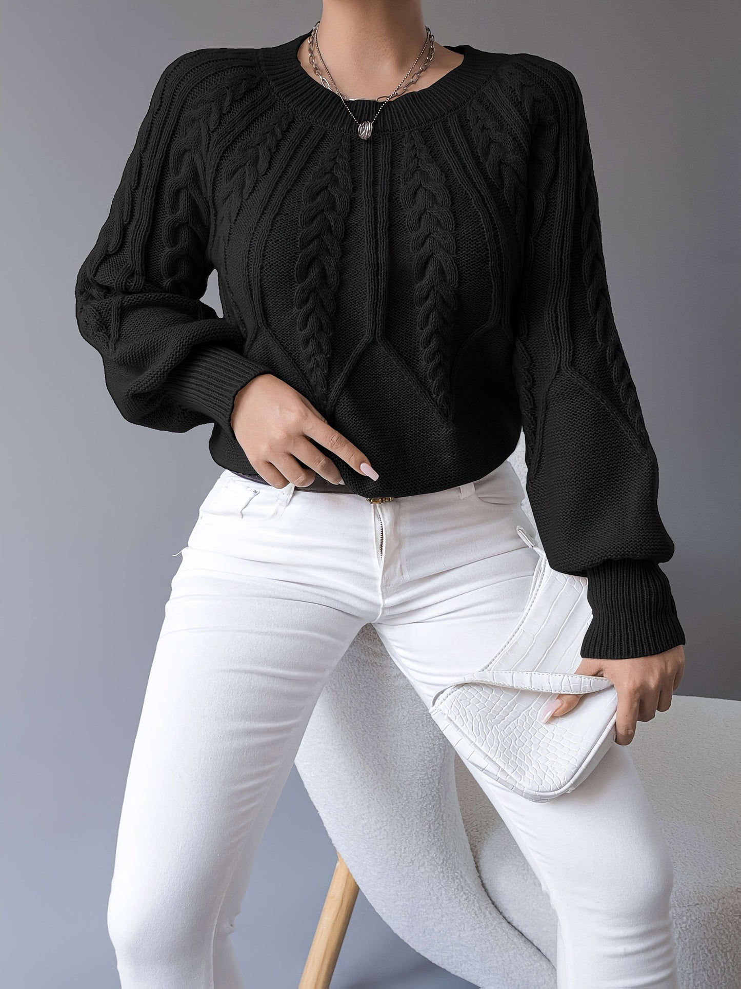 Kvinde iført sort strikket sweater med kabelstrik og ballonærmer, stylet med hvide bukser og halskæde, fotograferet forfra_8