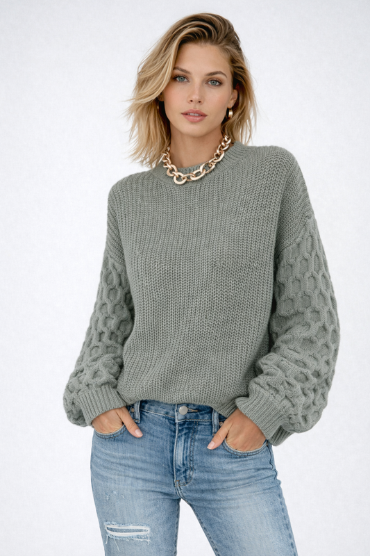 Model iført en grå strikket sweater med ballonærmer og kabelmønster sammen med lyse jeans og en guldfarvet kædehalskæde._0