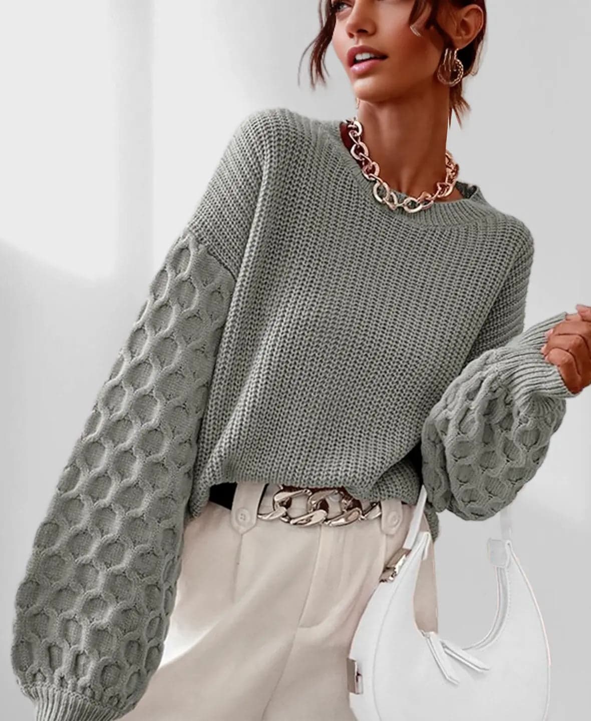 Model iført en grå strikket sweater med ballonærmer og kabelmønster sammen med lyse jeans og en guldfarvet kædehalskæde._1