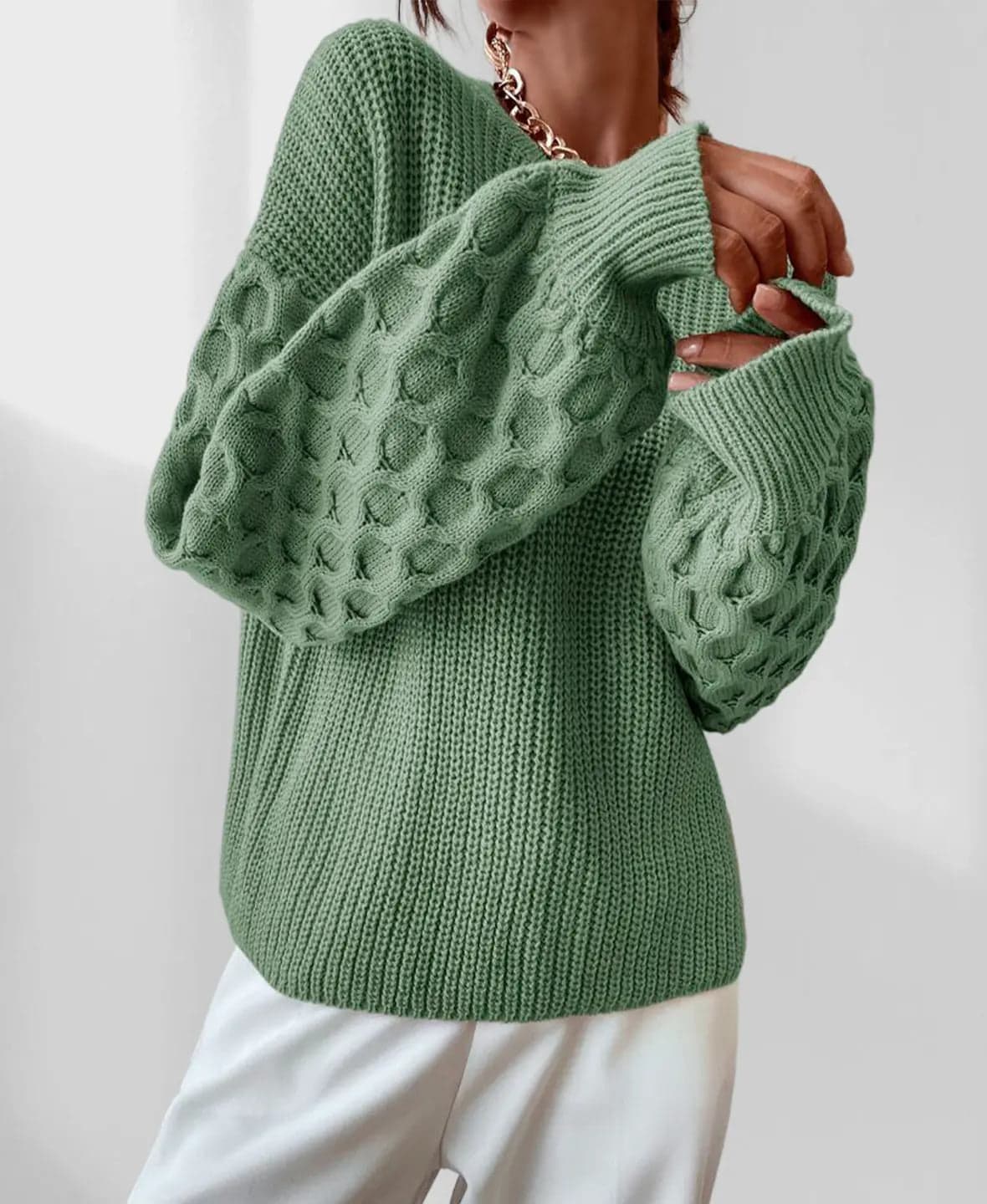 Model iført en grå strikket sweater med ballonærmer og kabelmønster sammen med lyse jeans og en guldfarvet kædehalskæde._13
